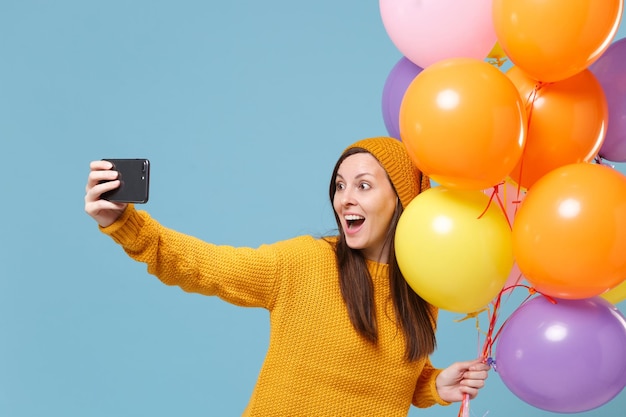 Mujer emocionada con sombrero de suéter posando aislada de fondo azul. Fiesta de cumpleaños, concepto de emociones de la gente. Simulacros de espacio de copia. Celebrando sostener globos aerostáticos haciendo selfie en el teléfono móvil.