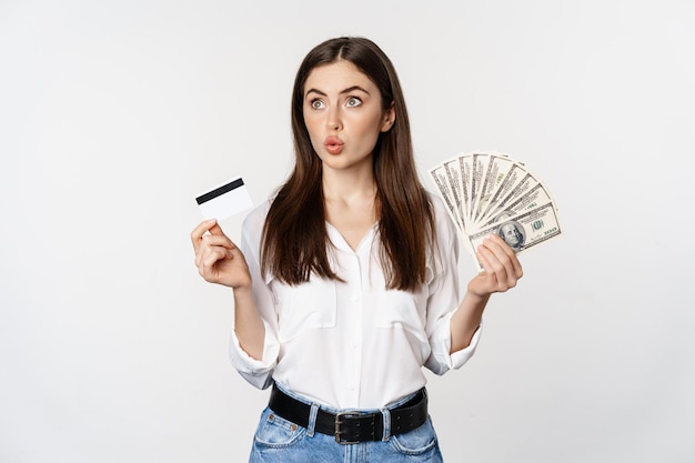 Mujer emocionada con tarjeta de crédito y dinero, mirando asombrada por el dinero en efectivo, de pie contra el fondo blanco