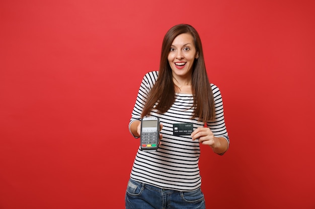 Mujer emocionada con terminal de pago bancario moderno inalámbrico para procesar y adquirir pagos con tarjeta de crédito, tarjeta negra aislada sobre fondo rojo. Personas sinceras emociones, estilo de vida. Simulacros de espacio de copia.