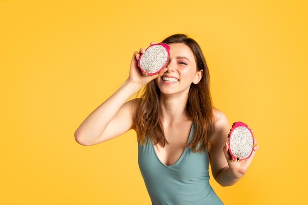 Mujer emocionada en traje de baño sosteniendo rodajas de pitaya cerca de la cara disfrutando de una sabrosa fruta jugosa de pie en