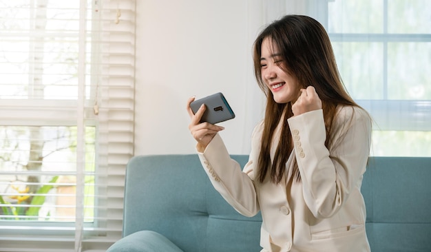 Una mujer emocionada usa el teléfono móvil levantando el puño para celebrar la victoria en línea o recibió buenas noticias en el sofá de la sala de estar en casa, una mujer asiática feliz divertida en un teléfono inteligente sintiéndose sorprendida emocionada