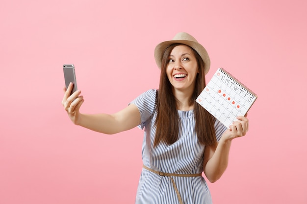 Mujer emocionada en vestido azul haciendo selfie en teléfono móvil, manteniendo el calendario de períodos para comprobar los días de menstruación aislados sobre fondo rosa. Concepto médico, sanitario, ginecológico. Copie el espacio.
