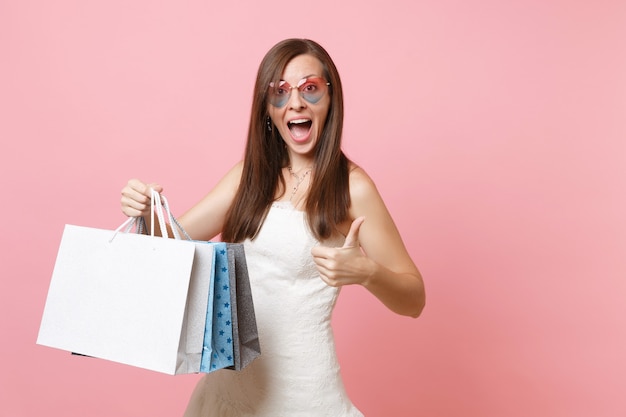 Mujer emocionada en vestido blanco, gafas de corazón mostrando el pulgar hacia arriba sosteniendo bolsas de paquetes multicolores con compras después de ir de compras