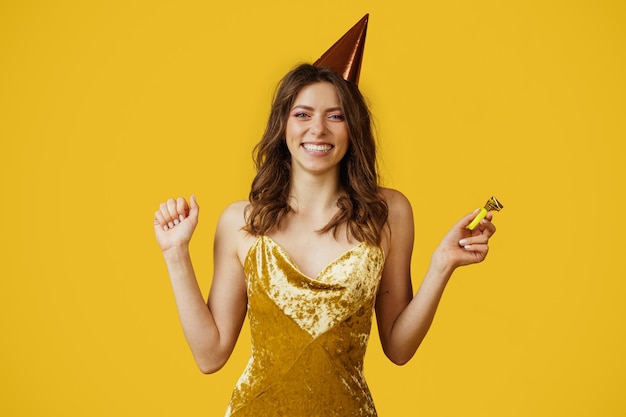 Mujer emocionada con vestido y gorra de cumpleaños sosteniendo cuerno de fiesta celebrando su día especial en el fondo amarillo del estudio