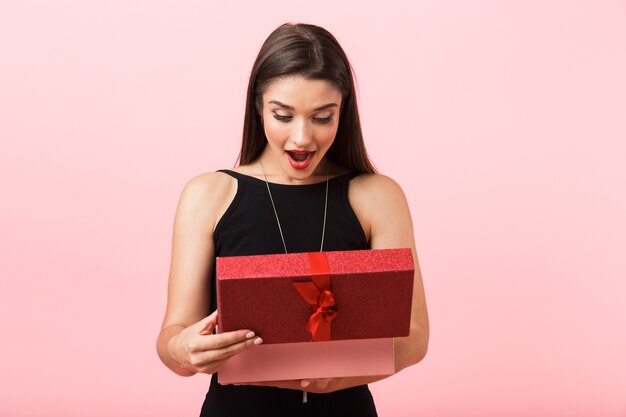 Mujer emocionada con vestido negro sosteniendo caja de regalo abierta aislada sobre fondo rosa