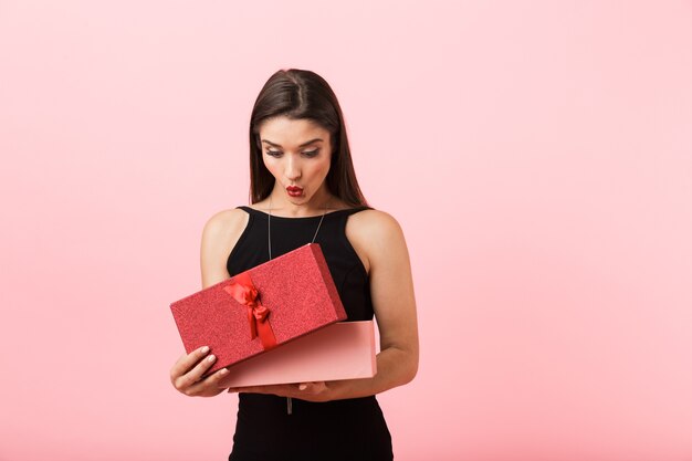 Mujer emocionada con vestido negro sosteniendo caja de regalo abierta aislada sobre fondo rosa