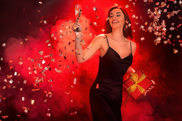Mujer emocionada en vestido negro sostiene la caja roja de regalo y una copa de champán concepto de vacaciones de tiempo de fiesta