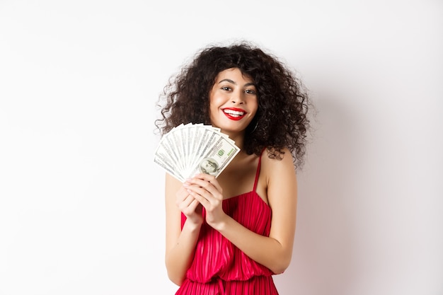 Mujer emocionada en vestido rojo ganando dinero, mostrando billetes de un dólar y sonriendo feliz, de pie sobre fondo blanco.