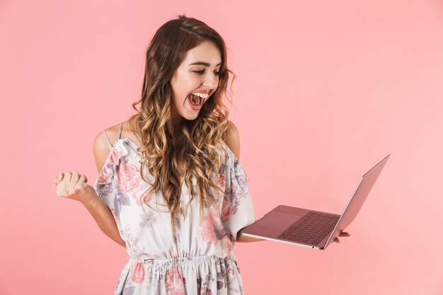 Mujer emocionada en vestido sosteniendo y usando portátil plateado, aislado en rosa