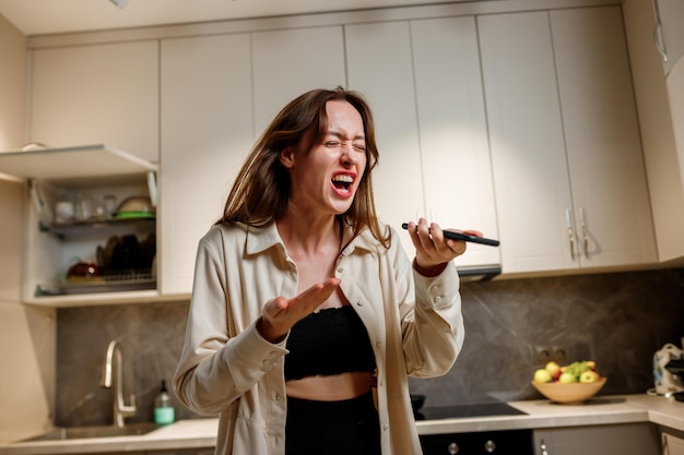 Mujer emocional enojada gritando expresivamente al teléfono inteligente mientras responde una llamada telefónica o graba un mensaje de voz mientras cocina en la cocina moderna