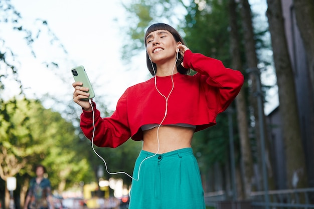 Foto mujer emocional escuchando música en la calle, linda mujer milenaria en un elegante suéter rojo con auriculares y escuchar música