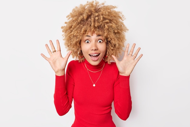 Mujer emocional positivamente sorprendida con cabello rizado levanta las manos reacciona ante algo increíble no puede creer que sus ojos reciban un regalo inesperado viste cuello alto rojo aislado en la pared blanca