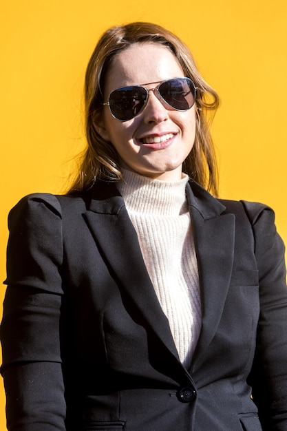 Mujer empresaria ejecutiva con chaqueta negra y suéter de lana y gafas de sol sobre fondo amarillo en la calle con espacio para copiar mirando a la cámara y sonriendo Concepto de mujer empresaria