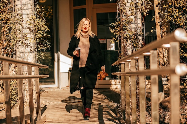 Mujer empresaria segura de sí misma va a tomar un café en el parque de otoño de la ciudad en un día soleado, obtiene satisfacción de una taza de té caliente y aire picante de otoño. Estilo de vida. La vida en el momento. Foto de cuerpo entero.