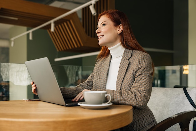 Mujer empresaria sentada en una mesa con una computadora portátil Mujer pelirroja trabajando de forma remota desde un café