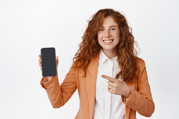 Mujer empresaria sonriente señalando con el dedo el teléfono móvil que se muestra en el teléfono inteligente de pie en traje contra el fondo blanco