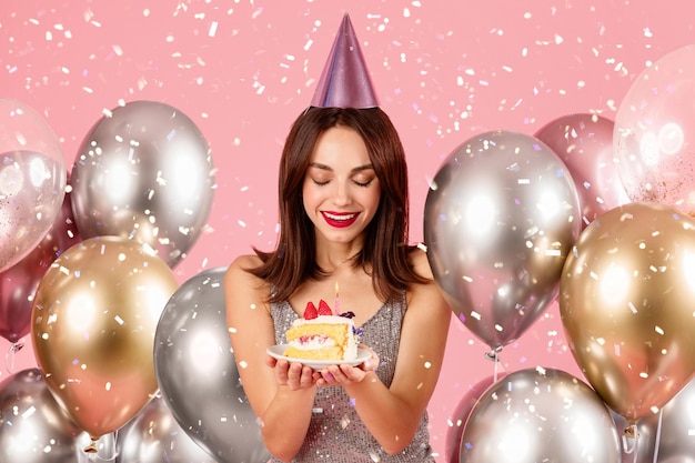 Foto mujer encantada con un sombrero festivo y un vestido brillante admirando una rebanada de pastel de cumpleaños