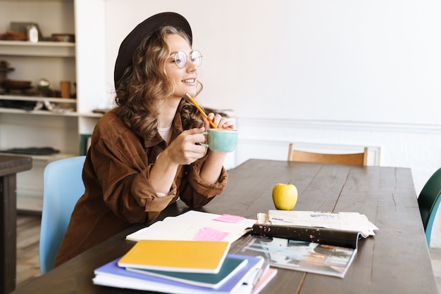 Mujer encantadora alegre en anteojos tomando café mientras estudia con libros de ejercicios en casa