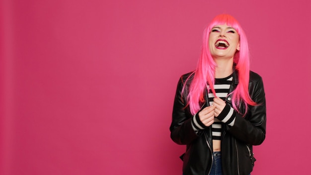 Mujer encantadora alegre con peluca de pelo rosa riendo y divirtiéndose en la cámara, mostrando una sonrisa sincera en el estudio. Joven feliz con chaqueta de cuero disfrutando de bromas y riéndose.