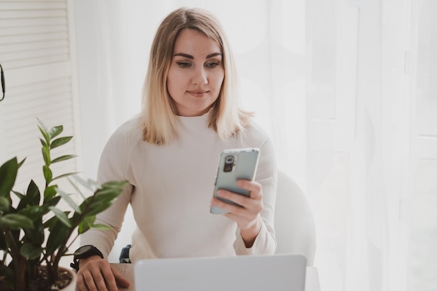 Una mujer encantadora y alegre que usa una computadora portátil y un teléfono móvil sonriendo mientras se sienta en el lugar de trabajo