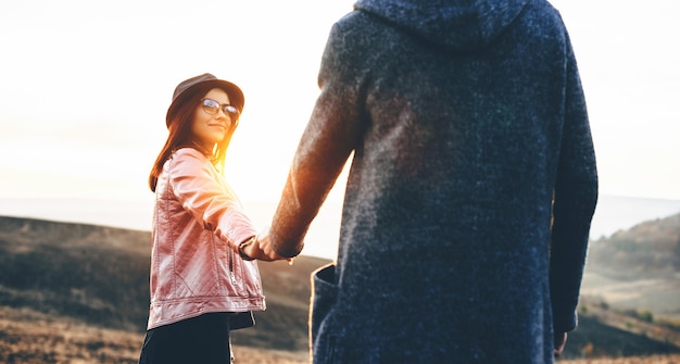 Mujer encantadora con anteojos y sombrero sosteniendo la mano de su amante y posando en un campo