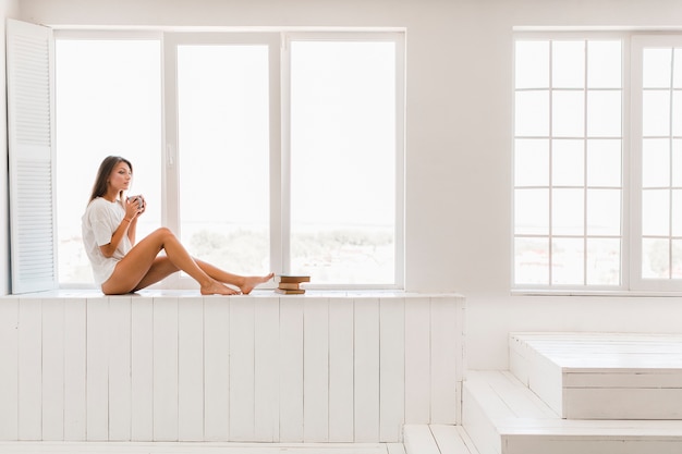 Mujer encantadora con bebida sentada junto a la ventana