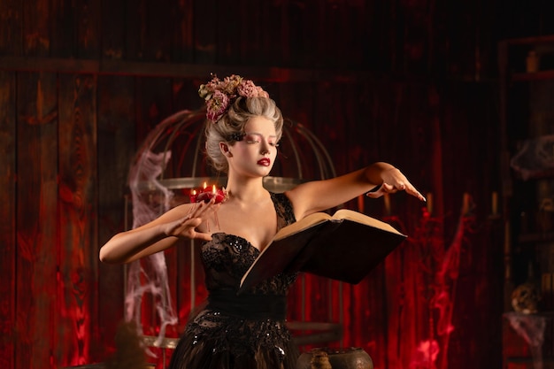 Mujer encantadora bruja flores en el pelo vestido negro vintage con libro de hechizos en el antiguo castillo Celebración de Halloween