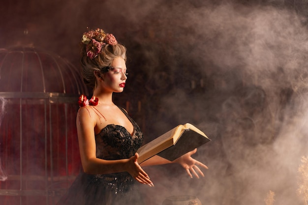 Mujer encantadora bruja flores en el pelo vestido negro vintage con libro de hechizos en el antiguo castillo Celebración de Halloween