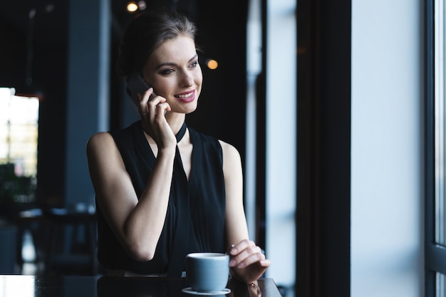 Mujer encantadora joven que llama con teléfono celular mientras está sentado en la cafetería durante el tiempo libre, mujer atractiva que tiene conversación hablando con el teléfono móvil mientras descansa en la cafetería.