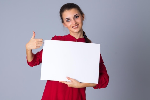 Mujer encantadora joven sosteniendo una hoja de papel blanca y pensando en un fondo gris en el estudio