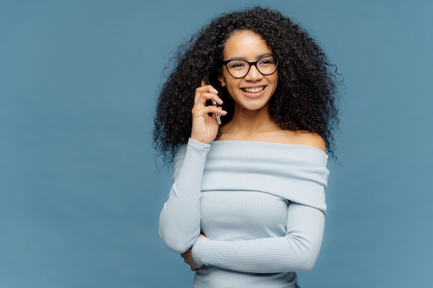 Mujer encantadora positiva tiene conversación telefónica, mantiene el teléfono móvil cerca del oído