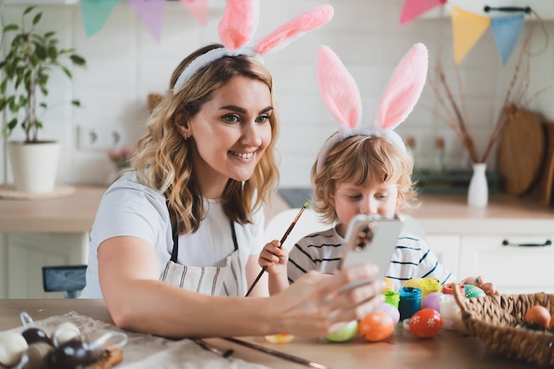 Una mujer encantadora y su hijo de dos años con orejas de conejo pintan huevos de Pascua con pinturas sentados en la mesa de la cocina