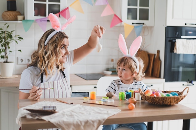 Una mujer encantadora y su hijo de dos años con orejas de conejo pintan huevos de Pascua con pinturas sentados en la mesa de la cocina