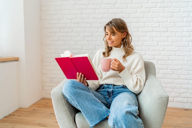 Mujer encantadora en suéter suave cálido disfrutando de una taza de té y un libro de lectura en la acogedora mañana de invierno