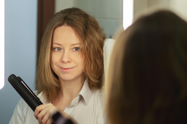 Una mujer enderezando el cabello en casa con un hierro para el pelo en el baño junto al espejo