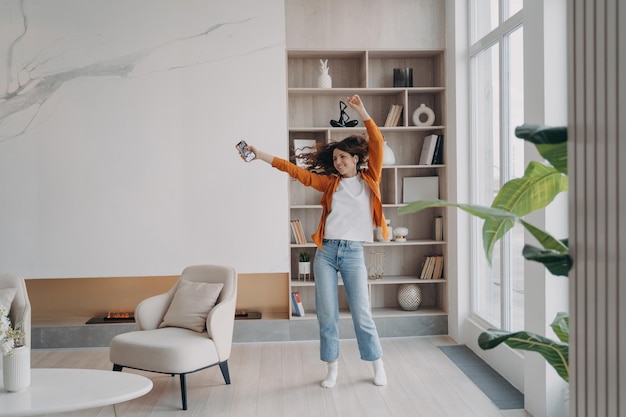 Mujer enérgica bailando en la sala de estar con música de teléfono inteligente en el moderno interior de un apartamento