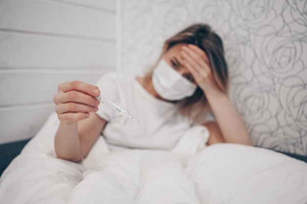 Mujer enferma acostada en la cama con termómetro y control de alta temperatura en el aislamiento de cuarentena en el hogar. Virus corona COVID-19. La infección causa enfermedad respiratoria primeros síntomas tos dolor de cabeza fiebre