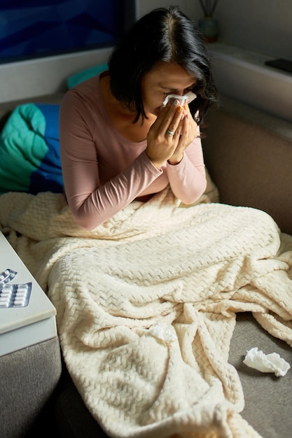 Mujer enferma con alergia a la gripe infecciones estacionales y secreción nasal limpiando la nariz, mujer enferma cubierta con una manta sentada en el sofá en casa.