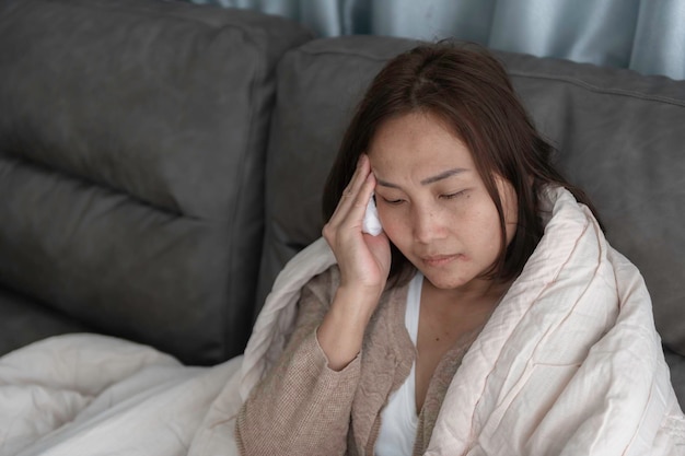 Una mujer enferma asiática se sienta en el sofá y se queda en casa. La mujer se sentía mal porque quería acostarse y descansar con fiebre alta.