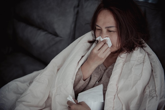 Una mujer enferma asiática se sienta en el sofá y se queda en casa. La mujer se sentía mal porque quería acostarse y descansar con fiebre alta.