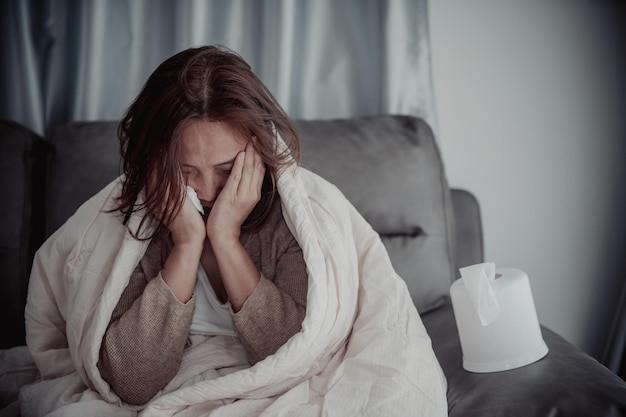 Una mujer enferma asiática se sienta en el sofá y se queda en casa. La mujer se sentía mal porque quería acostarse y descansar con fiebre alta.