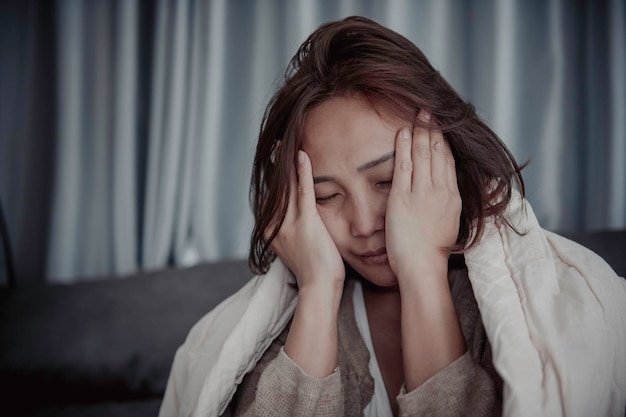 Una mujer enferma asiática se sienta en el sofá y se queda en casa. La mujer se sentía mal porque quería acostarse y descansar con fiebre alta.