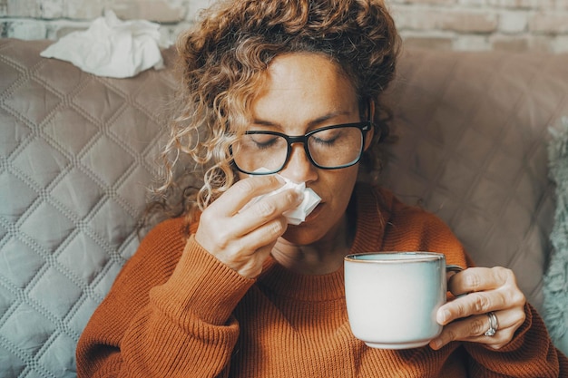 Mujer enferma en casa sonándose la nariz y cuidando la enfermedad del virus de la influenza Una mujer que usa papel tisú y bebe té de hierbas sola en casa Concepto de gripe resfriada en la temporada de invierno interior