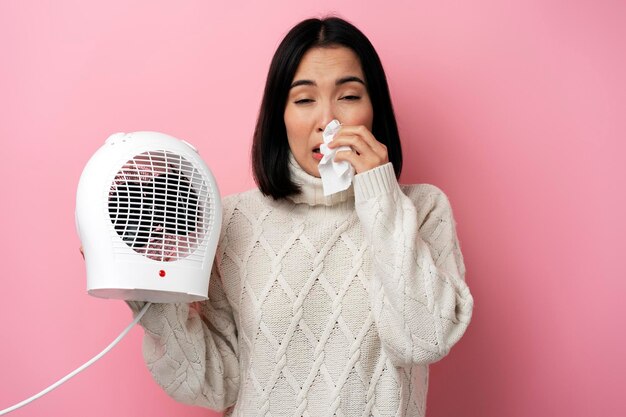 Mujer enferma estornudando en ropa de abrigo con radiador sobre fondo rosa