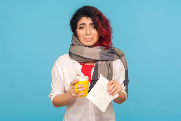 Mujer enferma y exhausta envuelta en una bufanda sosteniendo una taza de bebida caliente y una servilleta, mirando la cámara con una expresión enferma y miserable, sintiéndose mal, sufriendo influenza. foto de estudio, fondo azul