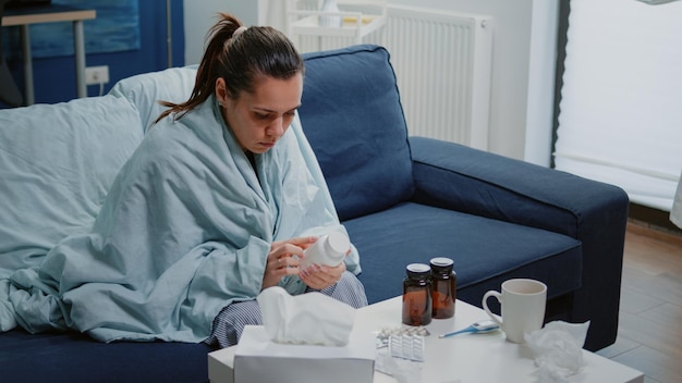 Mujer enferma leyendo la etiqueta del frasco de pastillas y medicamentos para curar los síntomas de la enfermedad. Adulto con frío sentado en una manta y sosteniendo un matraz con medicamentos y drogas contra la enfermedad