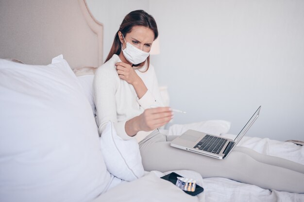 Mujer enferma en la máscara de protección facial acostado en la cama con un portátil con termómetro y pastillas en casa