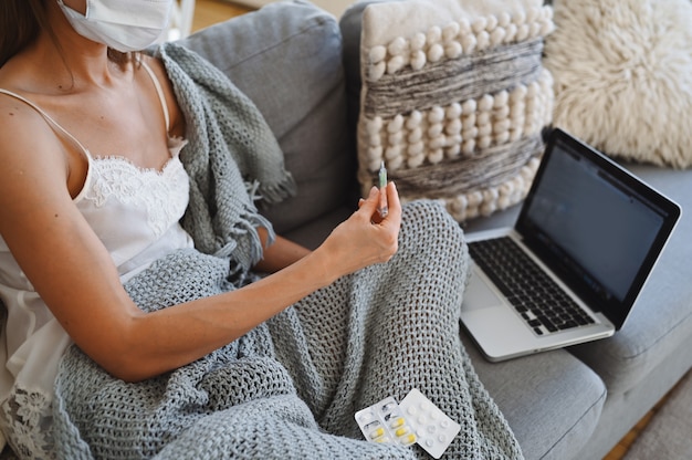 Mujer enferma con máscara de protección facial, sentado en el sofá con la computadora portátil y sosteniendo el termómetro y las píldoras durante la cuarentena casera auto aislamiento.