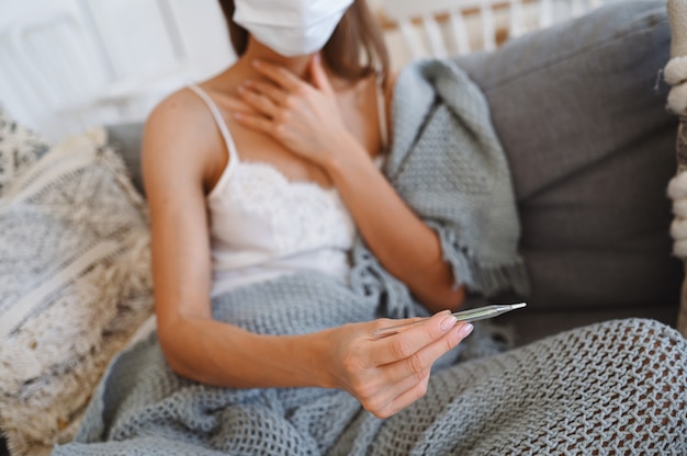 Mujer enferma con máscara de protección facial, sentado en el sofá con la computadora portátil y sosteniendo el termómetro y las píldoras durante la cuarentena casera auto aislamiento.