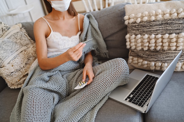 Mujer enferma con máscara de protección facial, sentado en el sofá con la computadora portátil y sosteniendo el termómetro y las píldoras durante la cuarentena casera auto aislamiento.